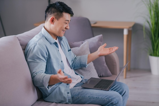 La foto di un uomo cinese divertente e positivo si siede sul divano e tiene il laptop mentre si gode una conversazione video remota indossa una maglietta casual in casa al chiuso