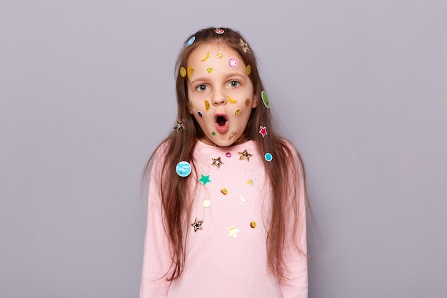 La foto di una bambina dai capelli castani scioccata e stupita coperta di adesivi in posa isolata su uno sfondo grigio tiene la bocca aperta vede enormi sconti nel negozio per bambini