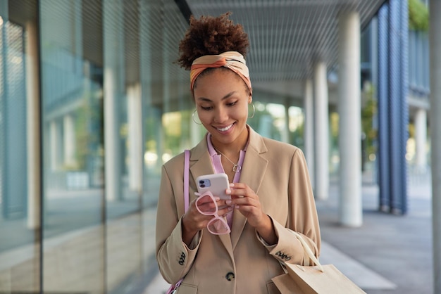 La foto di una donna dai capelli ricci alla moda indossa una giacca alla moda e la fascia per capelli usa il cellulare per chattare online