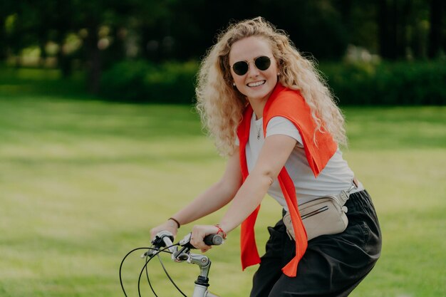 La foto di una donna europea riccia tiene le mani sul manubrio della bicicletta viaggia all'aperto nel parco indossa una maglietta casual e pantaloni neri sorride esprime positivamente la felicità Ricreazione della gente