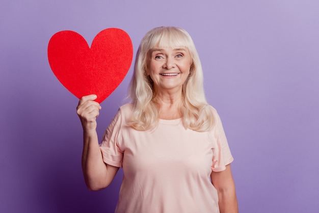 La foto di una donna graziosa e anziana sorridente tiene un grande cuore di cartellino rosso isolato su sfondo viola