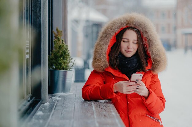 La foto di una giovane donna con una giacca rossa calda tiene un telefono cellulare moderno invia un messaggio di testo al fidanzato condivide le passioni sulle vacanze invernali si trova all'aperto durante la nevicata Concetto di tecnologia