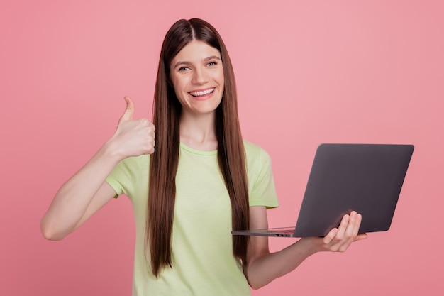 La foto di una giovane ragazza attraente felice e sorridente che usa il computer portatile mostra il pollice in su perfetto isolato su uno sfondo di colore rosa pastello