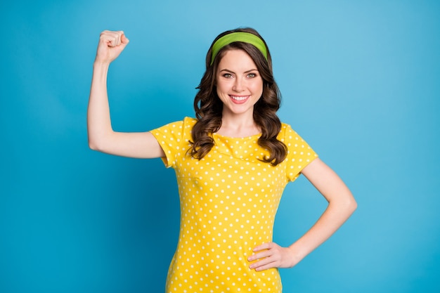 La foto di una ragazza allegra e positiva mostra i muscoli della mano che indossano abiti a pois isolati su uno sfondo di colore blu