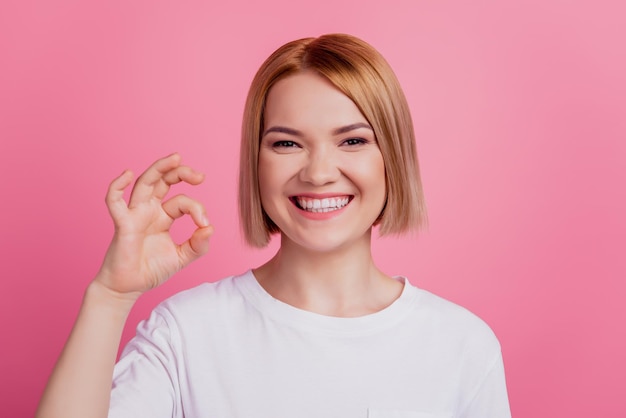 La foto di una signora bionda affascinante e positiva affidabile mostra il segno giusto indossa una maglietta bianca isolata su sfondo rosa