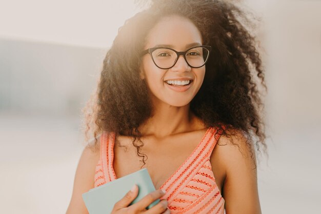 La foto di uno studente afroamericano spensierato in abito estivo tiene il libro si prepara per l'esame passeggiate attraverso le strade della città gode di una calda giornata di sole ha un sorriso affascinante sul viso Concetto di etnia della gente