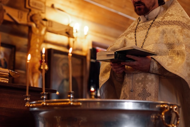 La foto ritagliata del sacerdote indossa la tonaca cerimoniale in piedi al font decorato con candele accese che leggono il libro di preghiera