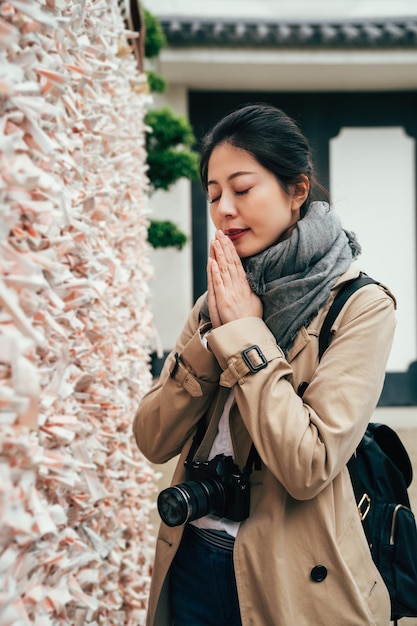 la fotografa asiatica sperimenta la cultura tradizionale giapponese. la ragazza mette insieme i palmi delle mani pregando aggraziando vicino al muro dello scivolo della fortuna. molti fogli con desideri e speranze appesi alla cravatta.