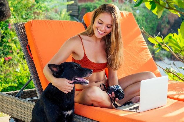 La fotografa surfista freelance della donna sta viaggiando con la sua ragazza freelancer doghappy che fa surf in un elegante costume da bagno bikini corallo che lavora con un computer portatile sulla spiaggia in un resort sul lettino arancione