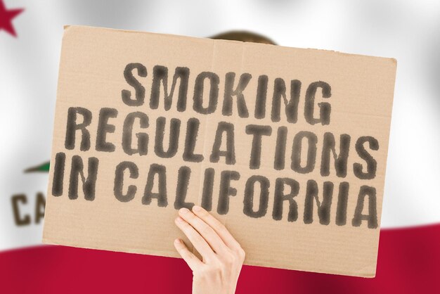 La frase "Norme sul fumo in California" su uno striscione in mano da uomo con bandiera della California sfocata sullo sfondo. giudiziario. Fuori. Luogo. Inquinamento. illegale. Rifiuto. Negato