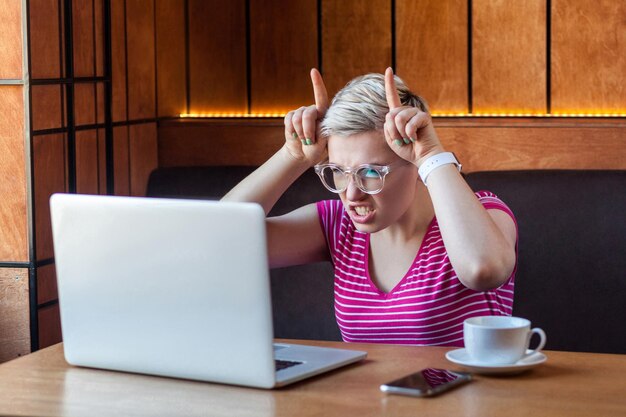 La freelance aggressiva arrabbiata della giovane ragazza con i capelli corti biondi in maglietta rosa e occhiali da vista è seduta al bar e di cattivo umore è un bullo con il dito che guarda il laptop al coperto