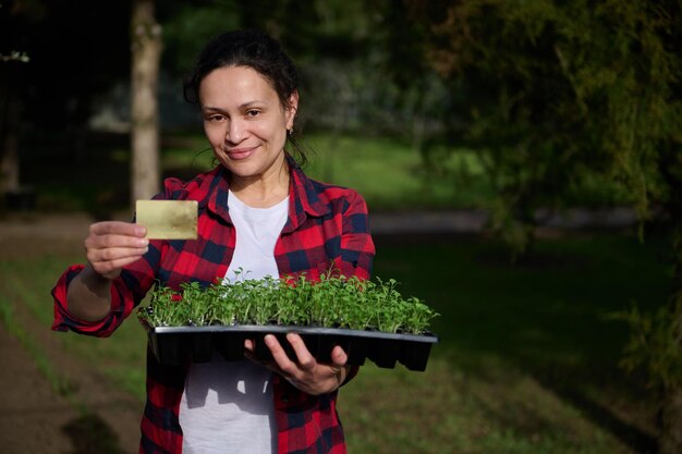 La giardiniera mostra una carta di credito dorata e tiene una cassetta con germogli crescenti di verdure culinarie su terreno nero in piedi nell'orto