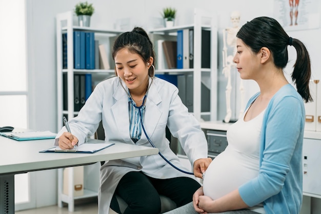 la ginecologa asiatica sta prendendo appunti e tiene uno stetoscopio vicino alla pancia per ascoltare il battito cardiaco del bambino mentre esamina la donna incinta in ospedale.