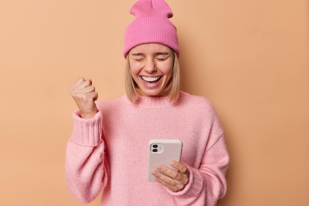 La gioiosa donna bionda stringe il pugno celebra il successo tiene il telefono cellulare di buon umore indossa un maglione rosa e un cappello isolato su sfondo beige Persone buona fortuna e concetto vincente