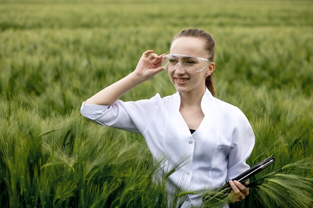 La giovane agricoltrice che indossa l'accappatoio bianco sta controllando i progressi del raccolto su un tablet nel campo di grano verde. Cresce un nuovo raccolto di grano. Concetto agricolo e di fattoria.