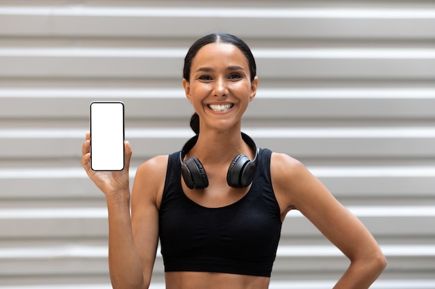La giovane atleta araba felice con le cuffie mostra lo smartphone con lo schermo vuoto sullo sfondo della parete