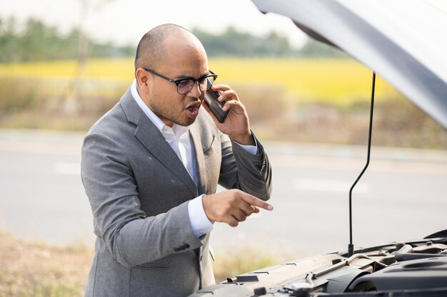 La giovane azienda asiatica ha rotto l'auto. Apre il cofano e controlla il motore. Usa lo smartphone per chiedere aiuto e servizio. Auto rotta sul ciglio della strada