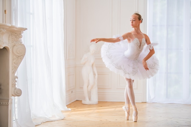 La giovane ballerina snella in un tutù bianco in scarpe da punta sta ballando in una grande e bella sala bianca.