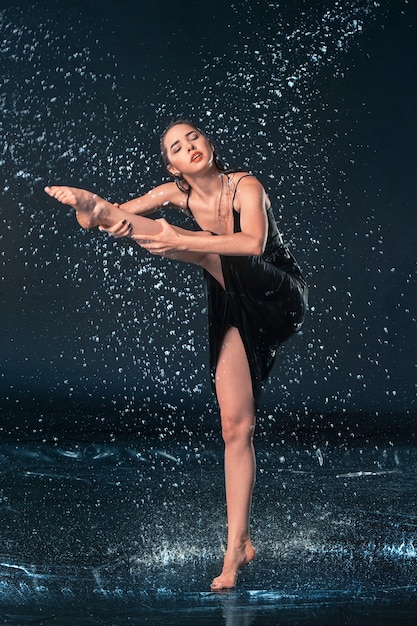 La giovane bella ballerina moderna che balla sotto le gocce d'acqua in studio blu aqua