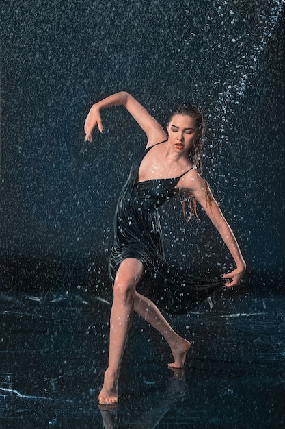 La giovane bella ballerina moderna che balla sotto le gocce d'acqua in studio blu aqua