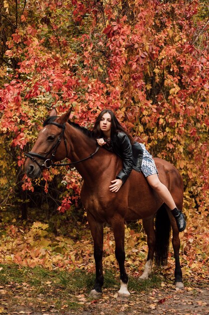 La giovane bella bruna con un vestito e una giacca di pelle nera è sexy sdraiata su un cavallo