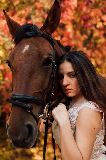 La giovane bella bruna in un vestito bianco abbraccia un cavallo marrone