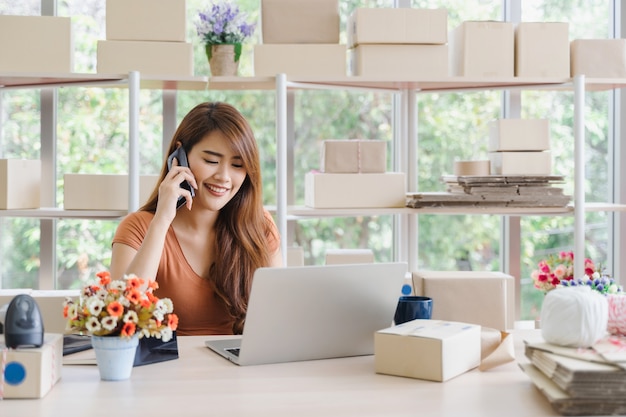 La giovane bella donna asiatica felice di affari nell&#39;abbigliamento casual con la faccina sta richiedendo riceve gli ordini del cliente con il computer portatile al suo ufficio domestico startup, concetto della PMI