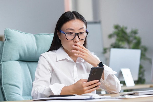 La giovane bella donna asiatica preoccupata di affari in vetri tiene il telefono nelle sue mani guarda