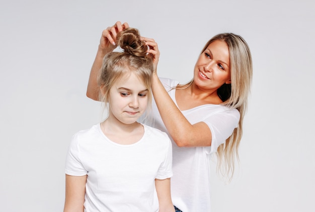 La giovane bella donna bionda fa lo stile di capelli per la ragazza. Svago comune di madre e figlia. Isolato su sfondo bianco