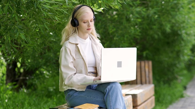 La giovane bella donna bionda in cuffie si siede su una panchina del parco con un computer portatile. Lavorare all'aperto in una calda giornata estiva. 4K UHD
