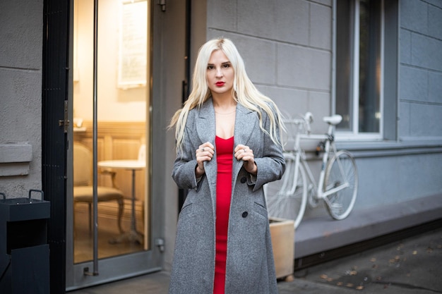 La giovane bella donna bionda in un cappotto di autunno sta su una via della città. Focalizzazione morbida. Concetto alla moda. Sfondo grigio