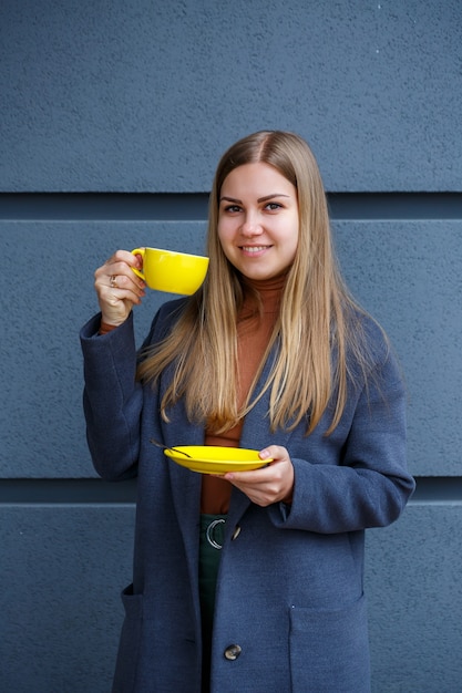 La giovane bella donna bionda in un cappotto grigio beve tè caldo da una tazza gialla in una fredda giornata autunnale. Deliziosa bevanda calda sulla terrazza del caffè