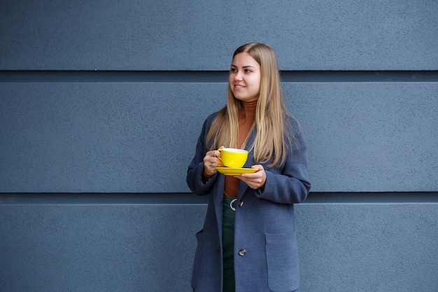 La giovane bella donna bionda in un cappotto grigio beve tè caldo da una tazza gialla in una fredda giornata autunnale. Deliziosa bevanda calda sulla terrazza del caffè