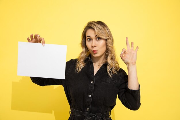 La giovane bella donna bionda in vestito nero tiene il foglio vuoto per l'iscrizione su fondo giallo in mani che indicano con le dita firmano ok, sorride e ride felicemente su fondo giallo in studio