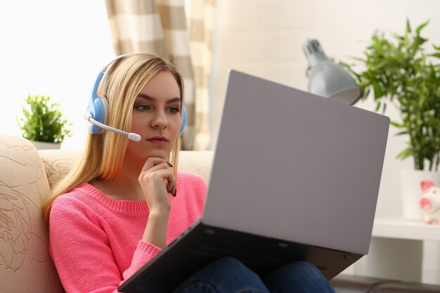 La giovane bella donna bionda si siede sul sofà in soggiorno tiene il computer portatile in armi ascolta la musica