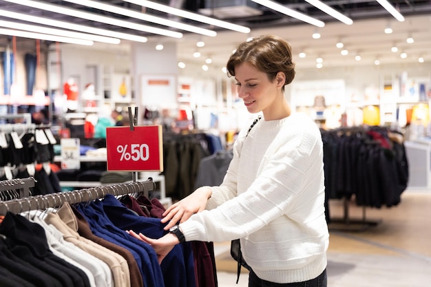 La giovane bella donna castana con un taglio di capelli corto in un maglione bianco sceglie i vestiti alla moda in un deposito in un centro commerciale