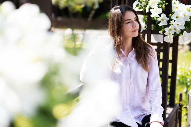 La giovane bella donna castana in camicia si siede sulla panca di legno con i fiori in giardino