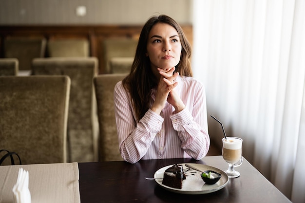 La giovane bella donna castana si siede nel ristorante del caffè della caffetteria all'interno con le dita attraversate