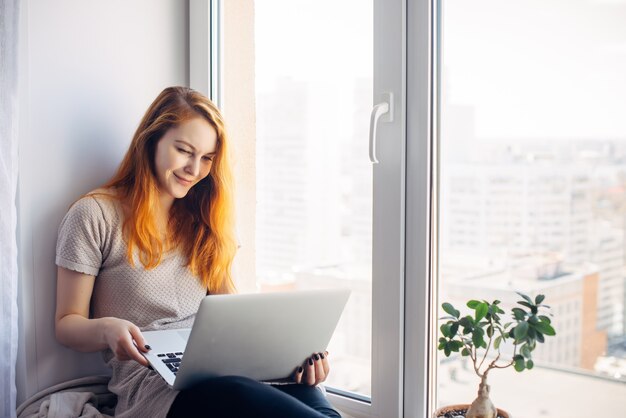 La giovane bella donna con capelli rossi lunghi si siede sul davanzale e guarda il computer portatile. Scrittrice con pc in grembo sullo sfondo della finestra in appartamento di città. Apprendimento online, concetto di freelance.