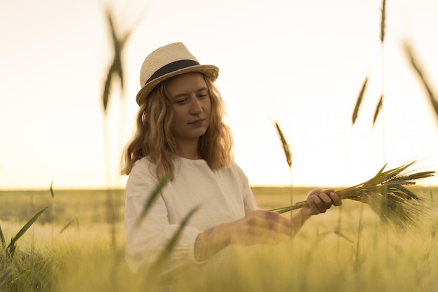 la giovane bella donna con i capelli lunghi biondi in un vestito bianco in un cappello di paglia raccoglie i fiori su un campo di grano. Capelli volanti al sole, estate. Tempo per sognatori, tramonto dorato.