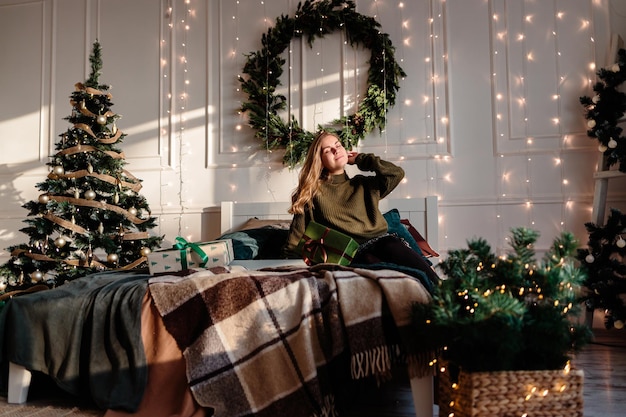 La giovane bella donna con un maglione in una stanza decorata per il Natale siede sullo sfondo di un albero di Natale