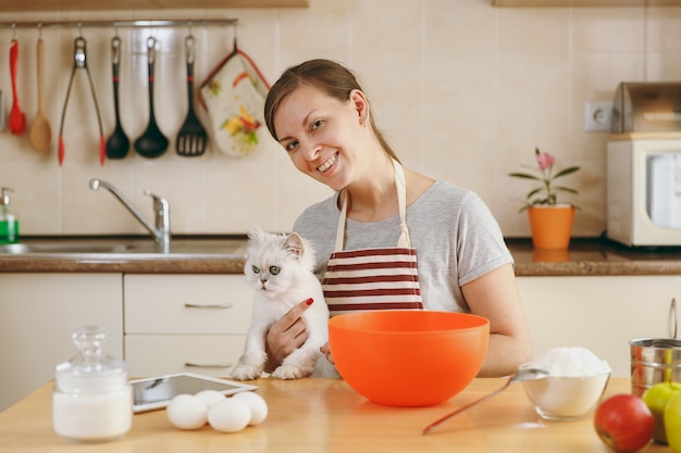 La giovane bella donna felice con un gatto persiano bianco prepara l'impasto per le torte con il tablet sul tavolo in cucina. Cucinare a casa. Prepara da mangiare.