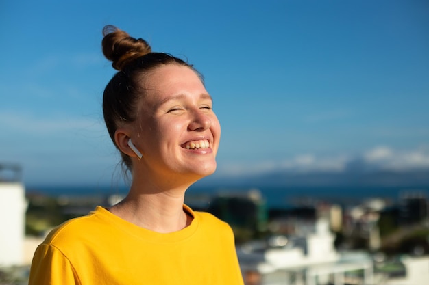 La giovane bella donna felice della ragazza positiva sta ascoltando la canzone di musica in auricolari senza fili medita alla giornata di sole di estate e sorride Concetto di libertà di felicità sullo sfondo naturale del cielo