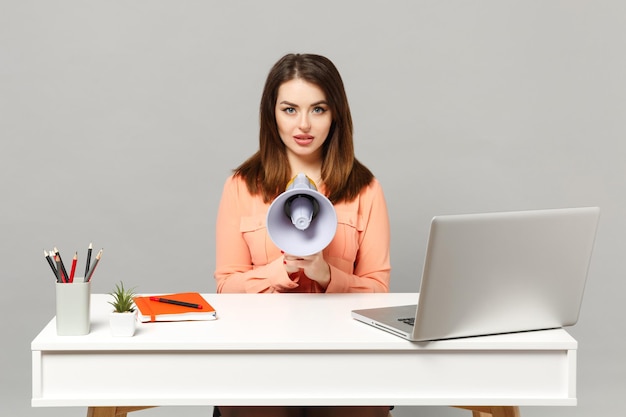 La giovane bella donna in abiti casual pastello che tiene il megafono si siede e lavora alla scrivania con un computer portatile isolato su sfondo grigio. Concetto di stile di vita di carriera aziendale di successo. Mock up spazio di copia.