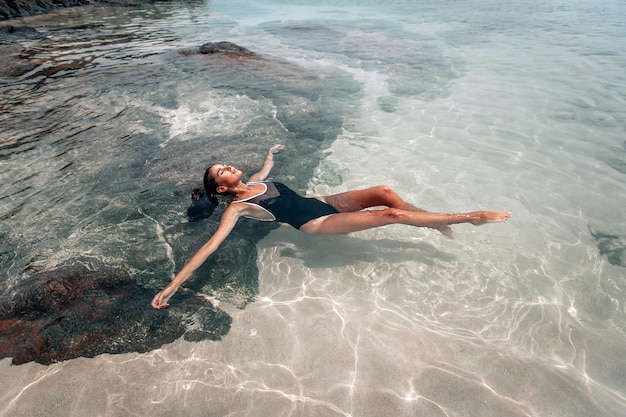 La giovane bella donna in costume da bagno nero si rilassa e prende il sole sdraiato nell'acqua sulla spiaggia. la vista dall'alto