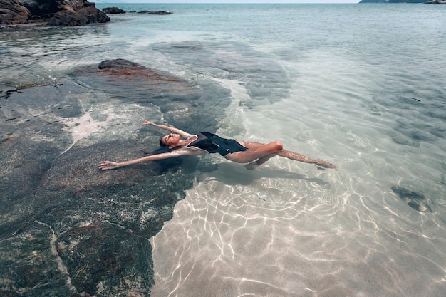 La giovane bella donna in costume da bagno nero si rilassa e prende il sole sdraiato nell'acqua sulla spiaggia. la vista dall'alto