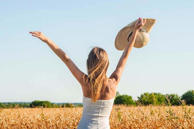 La giovane bella donna in un vestito bianco e un cappello solleva le sue mani sul campo