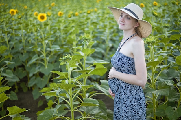 La giovane bella donna incinta si leva in piedi in un cappello e si veste sul campo dei girasoli di fioritura