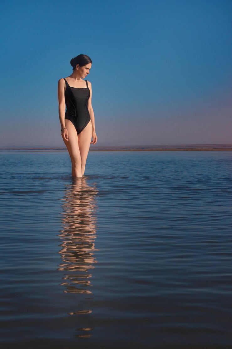 La giovane bella donna magra che indossa un costume da bagno nero sta nel mare e guarda il tramonto