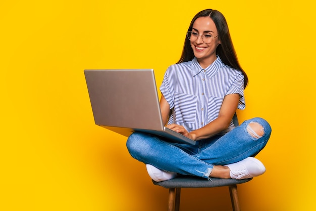 La giovane bella donna moderna è seduta sulla sedia con un computer portatile in mano mentre è isolata su sfondo giallo